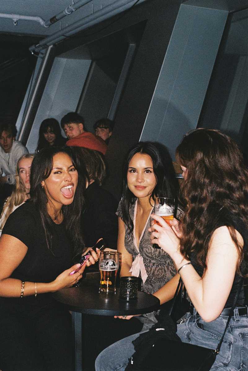 Three girls sit and stand around a small round table, each drinking a beer. One girl looks away from the camera, while the other two look directly at it, with the girl on the far left opening her mouth as if shouting. Many other bar patrons are visible in the background. Photograph.