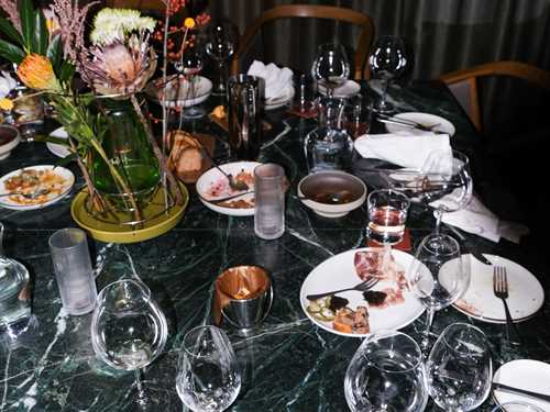 An abandoned restaurant table with used glasses and plates, along with discarded napkins and chairs pushed away from the table. Photograph.