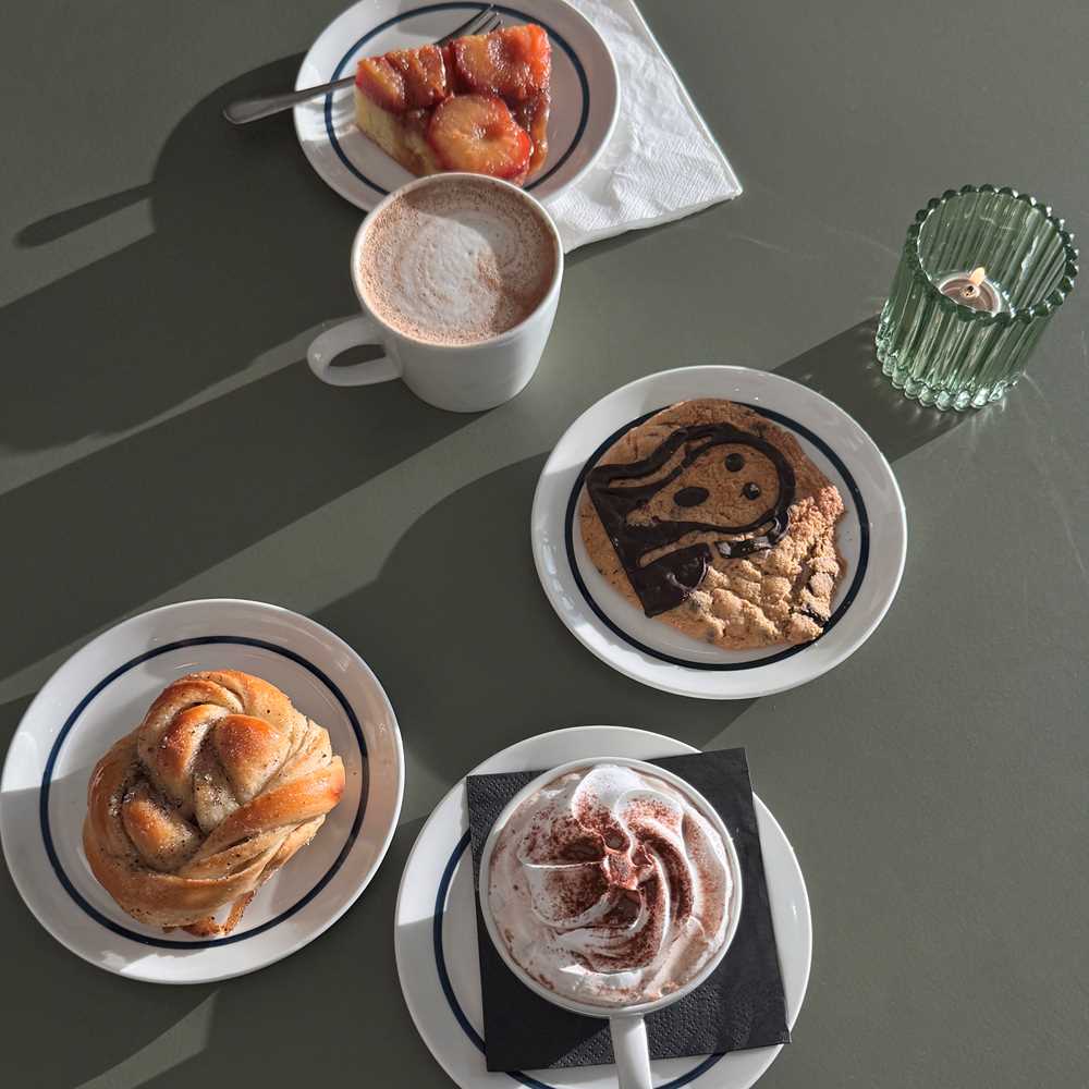 A green table seen from above, with a cup of mocha, a hot chocolate with cream, and three plates holding a cardamom bun, a cookie with The Scream motif, and a plum tart. Photograph.