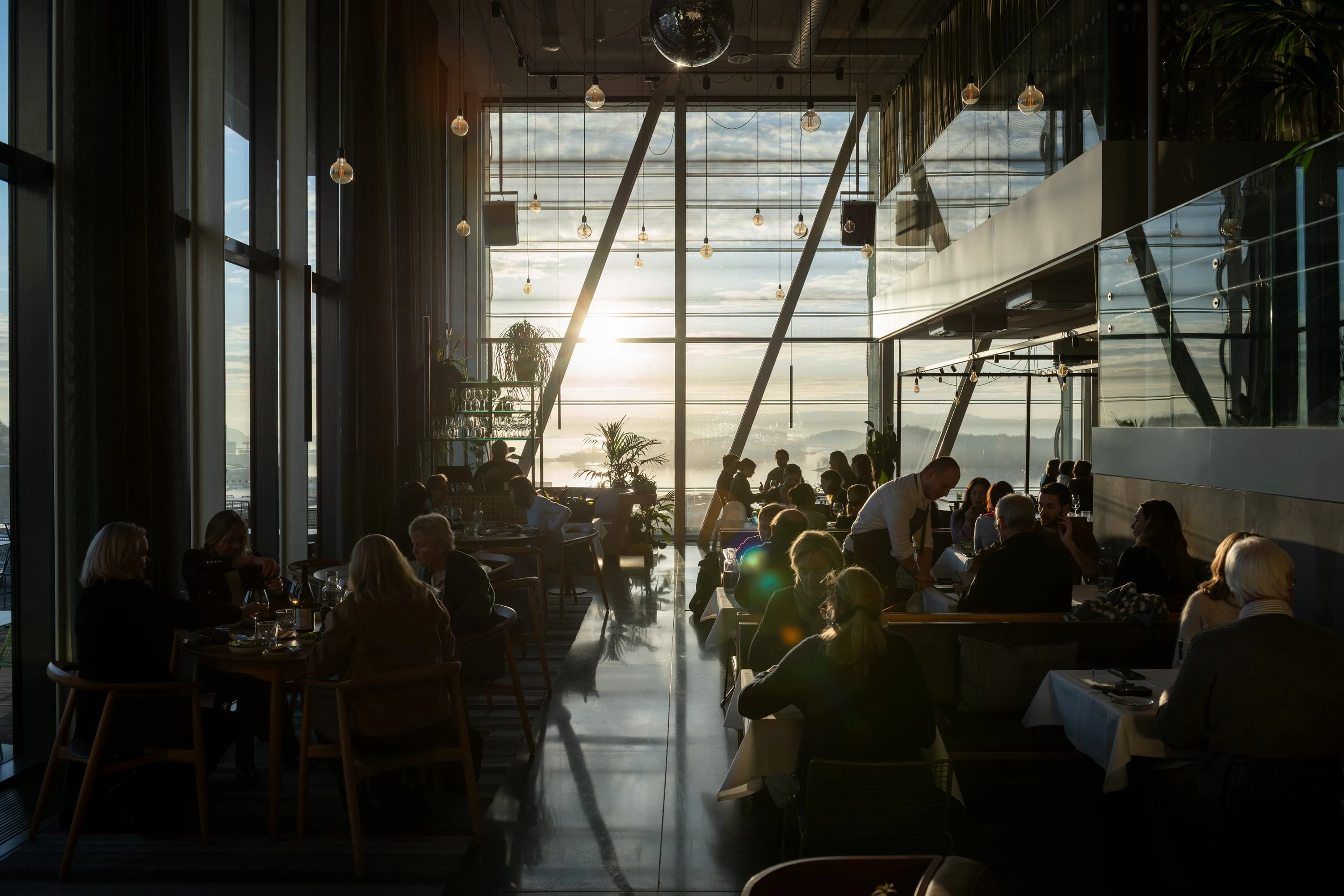 MUNCH restaurant bathed in sunlight streaming through the wall of windows at the end of the room. The restaurant is full, with people at every table and waiters moving between them. Photograph.
