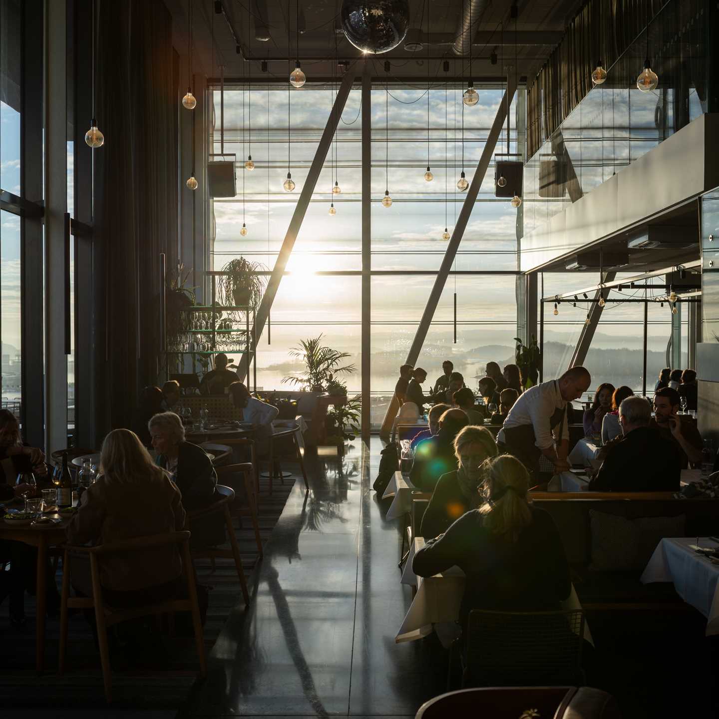 MUNCH restaurant bathed in sunlight streaming through the wall of windows at the end of the room. The restaurant is full, with people at every table and waiters moving between them. Photograph.