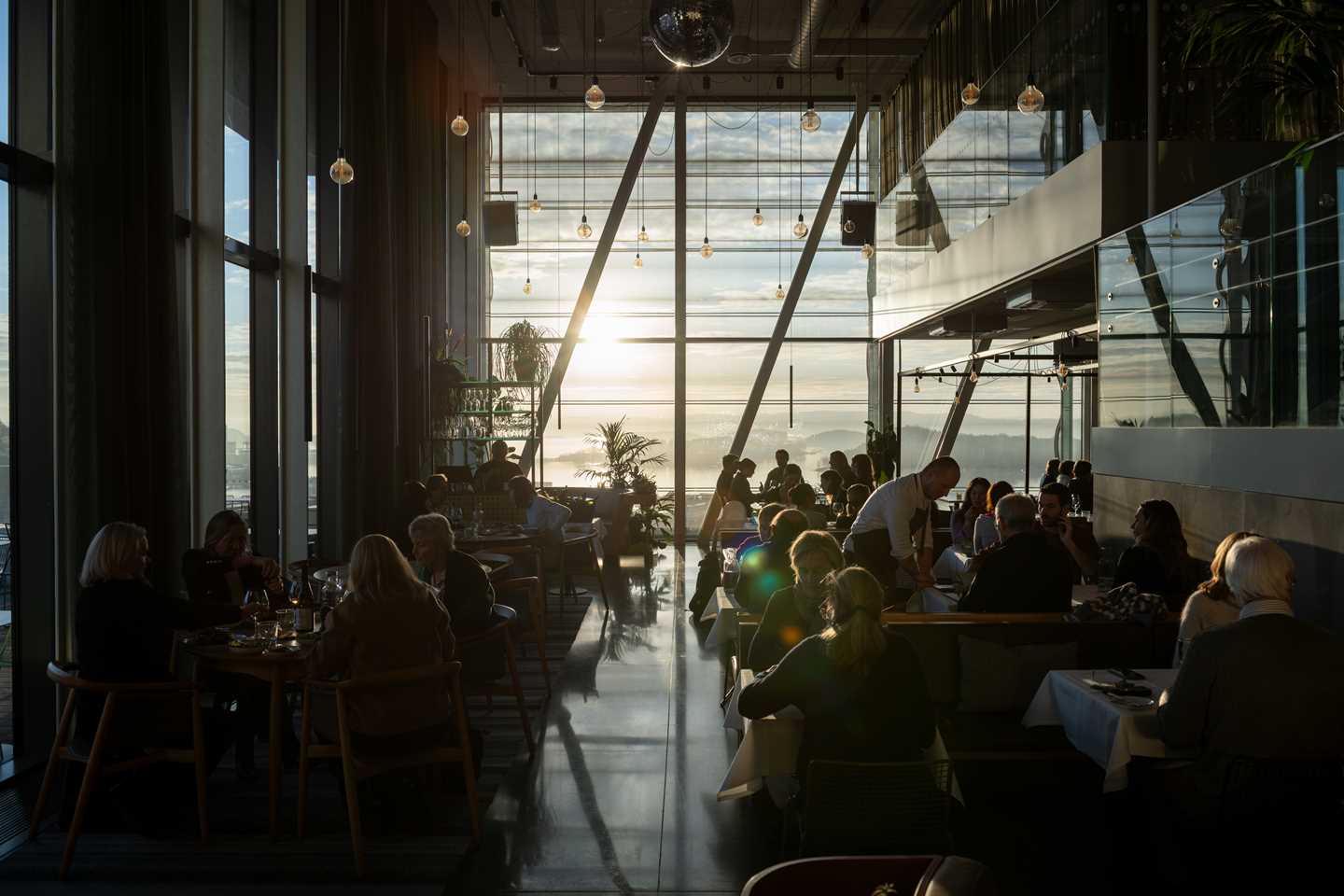 MUNCH restaurant bathed in sunlight streaming through the wall of windows at the end of the room. The restaurant is full, with people at every table and waiters moving between them. Photograph.