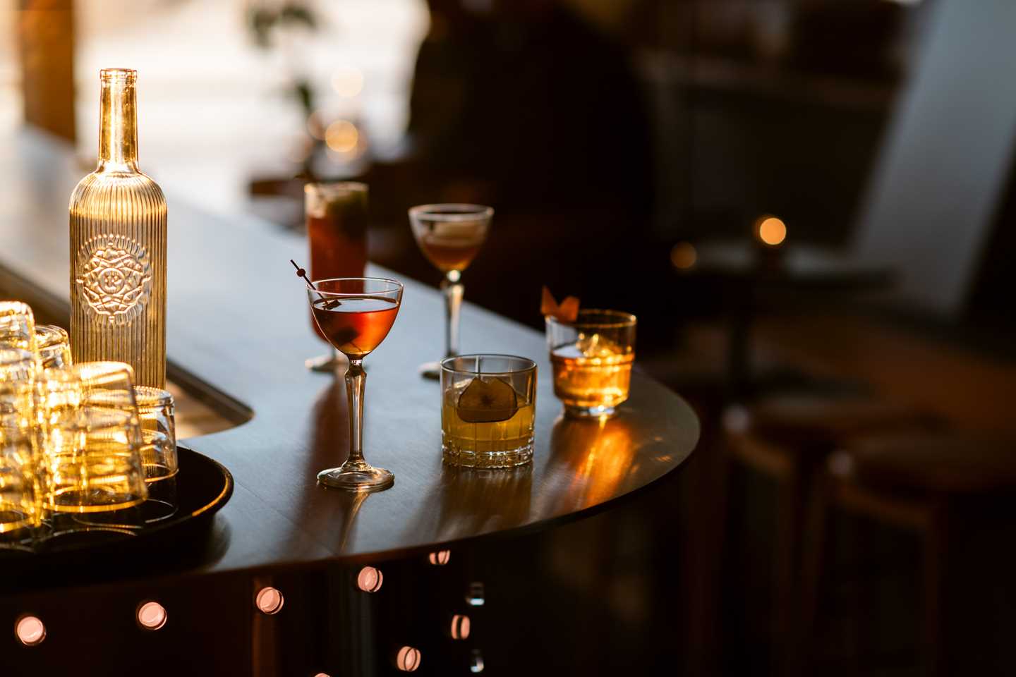 A bar counter with various drinks in different glasses. At the edge of the image, a tray holds empty glasses and a glass bottle filled with water. Photograph.