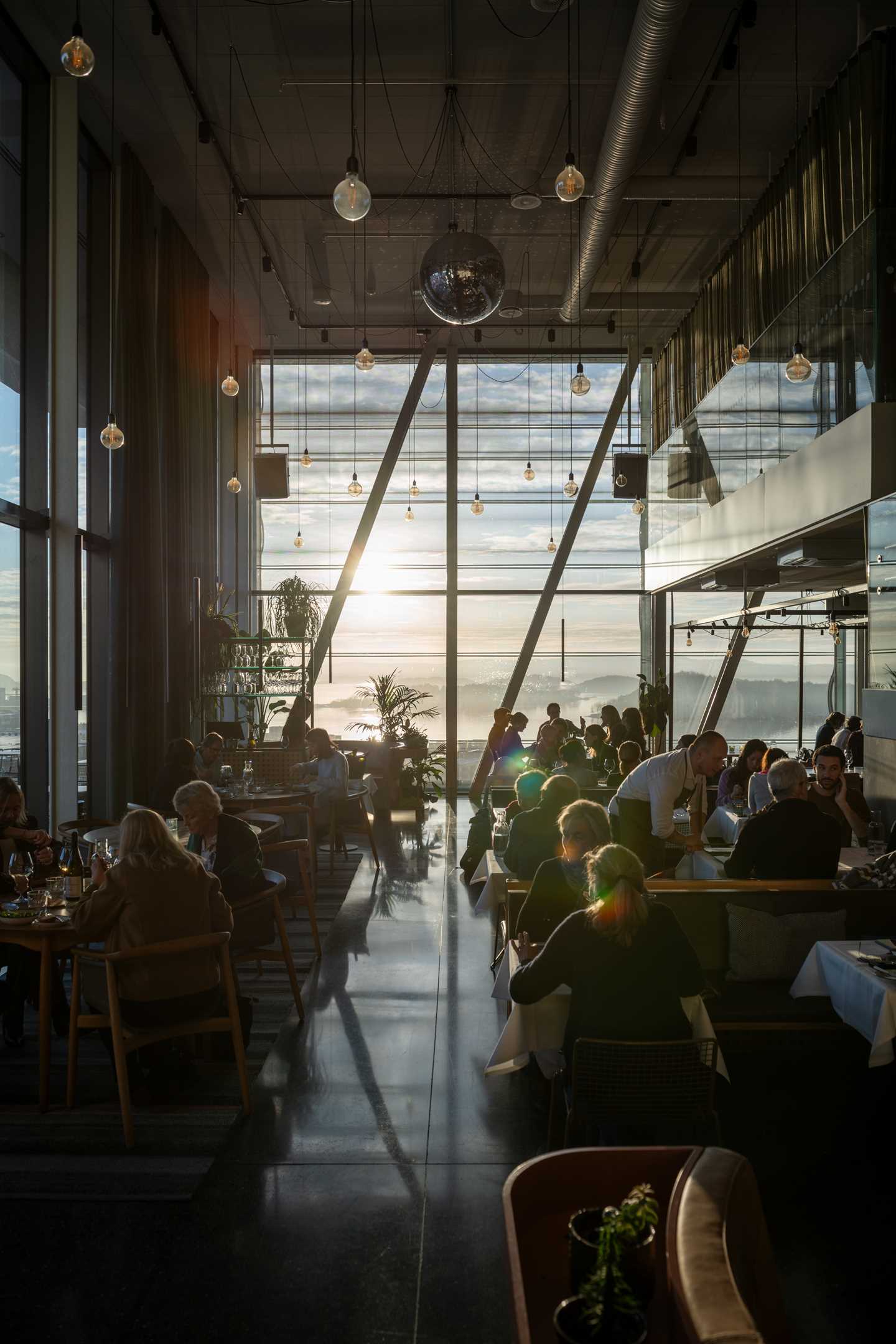 MUNCH’s restaurant on the top floor of the building. A full restaurant with people at every table and waiters talking to some of the guests. At the far end, floor-to-ceiling windows let in the sunlight, with a view over the Oslo Fjord. Photograph