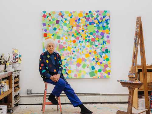 The artist Samia Halaby sits on a pink stool in her studio in front of one of her own works. The wall is white and the artwork consists of many colourful squares. To the right of the image stands an easel with paint stains, and to the left we see a shelving unit full of paint and painting materials. Photograph.