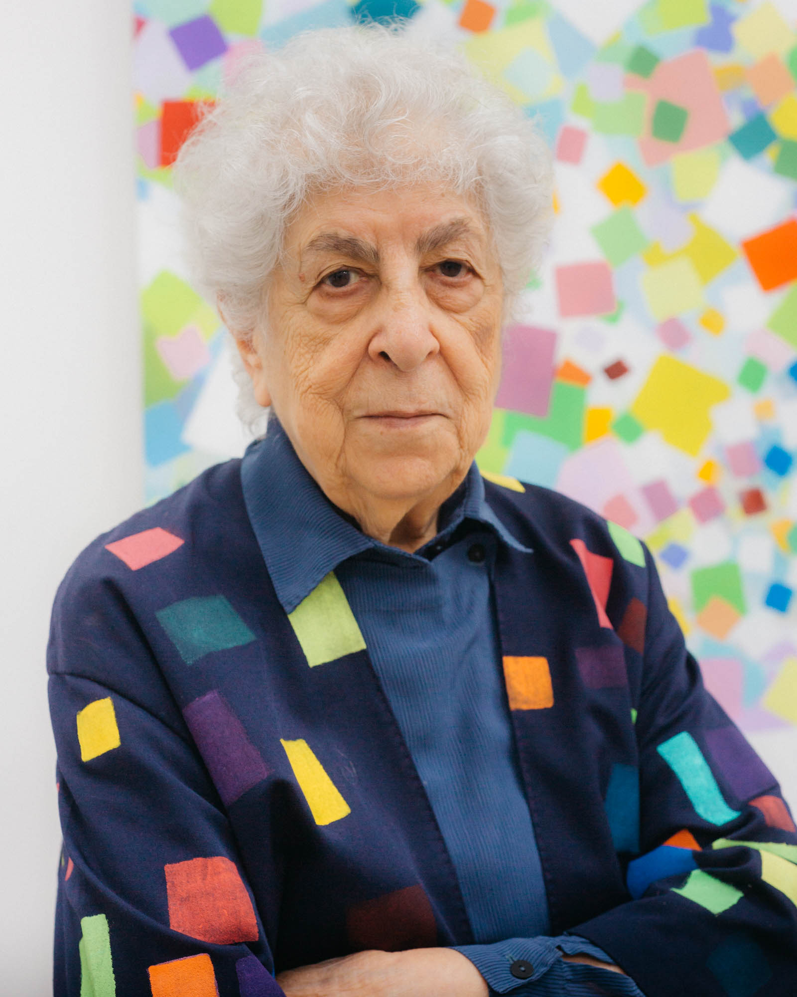 Portrait of the artist from the waist up, standing in front of one of her own paintings. Photography.