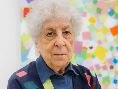 Portrait of the artist from the waist up, standing in front of one of her own paintings. Photography.