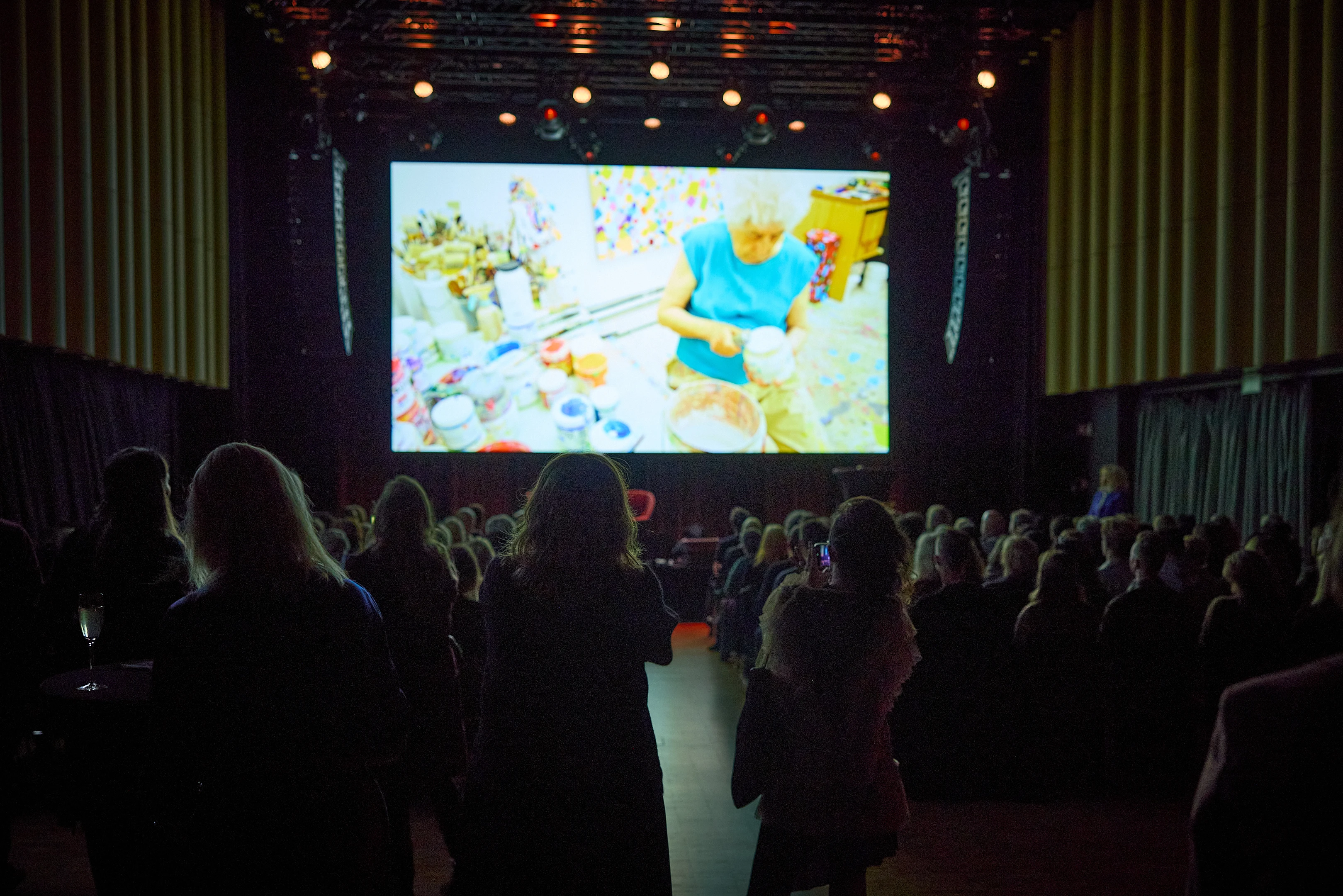 A packed banquet hall seen from the opposite side of the stage. All the rows of chairs are filled, and many people are standing in front of the camera. Everyone is watching a large screen behind the stage showing a film. Photograph.