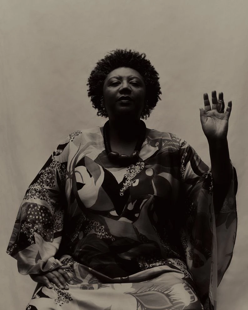 Portrait of a seated Rosana Paulino. She raises her left hand as if taking an oath. The image is in black and white. Photograph.
