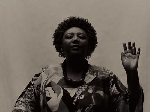 Portrait of a seated Rosana Paulino. She raises her left hand as if taking an oath. The image is in black and white. Photograph.
