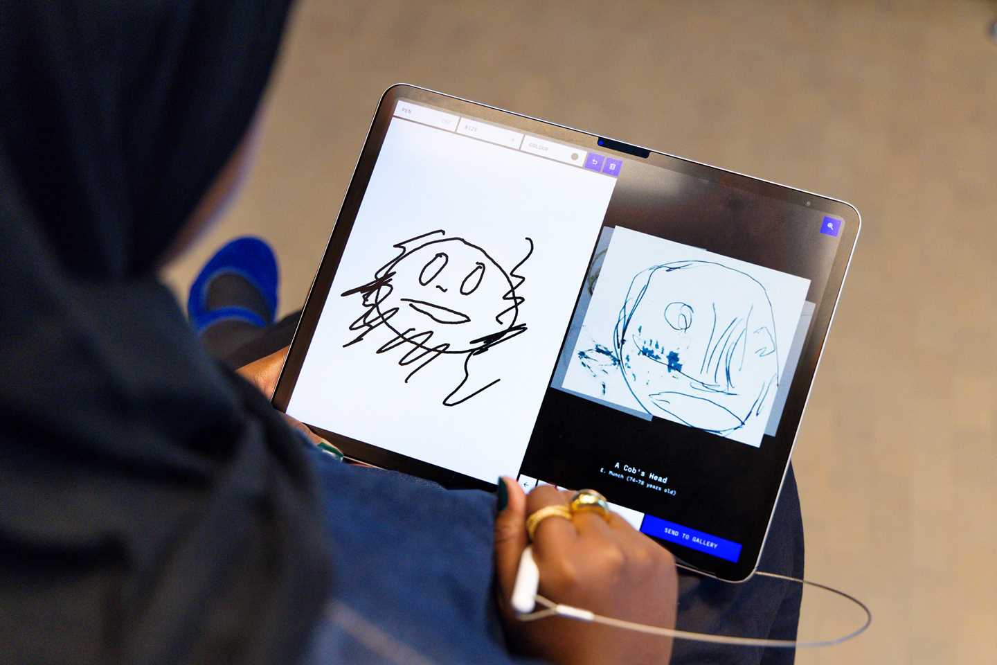 A woman is drawing on a tablet and is shown a similar drawing made by Edvard Munch. Photograph.