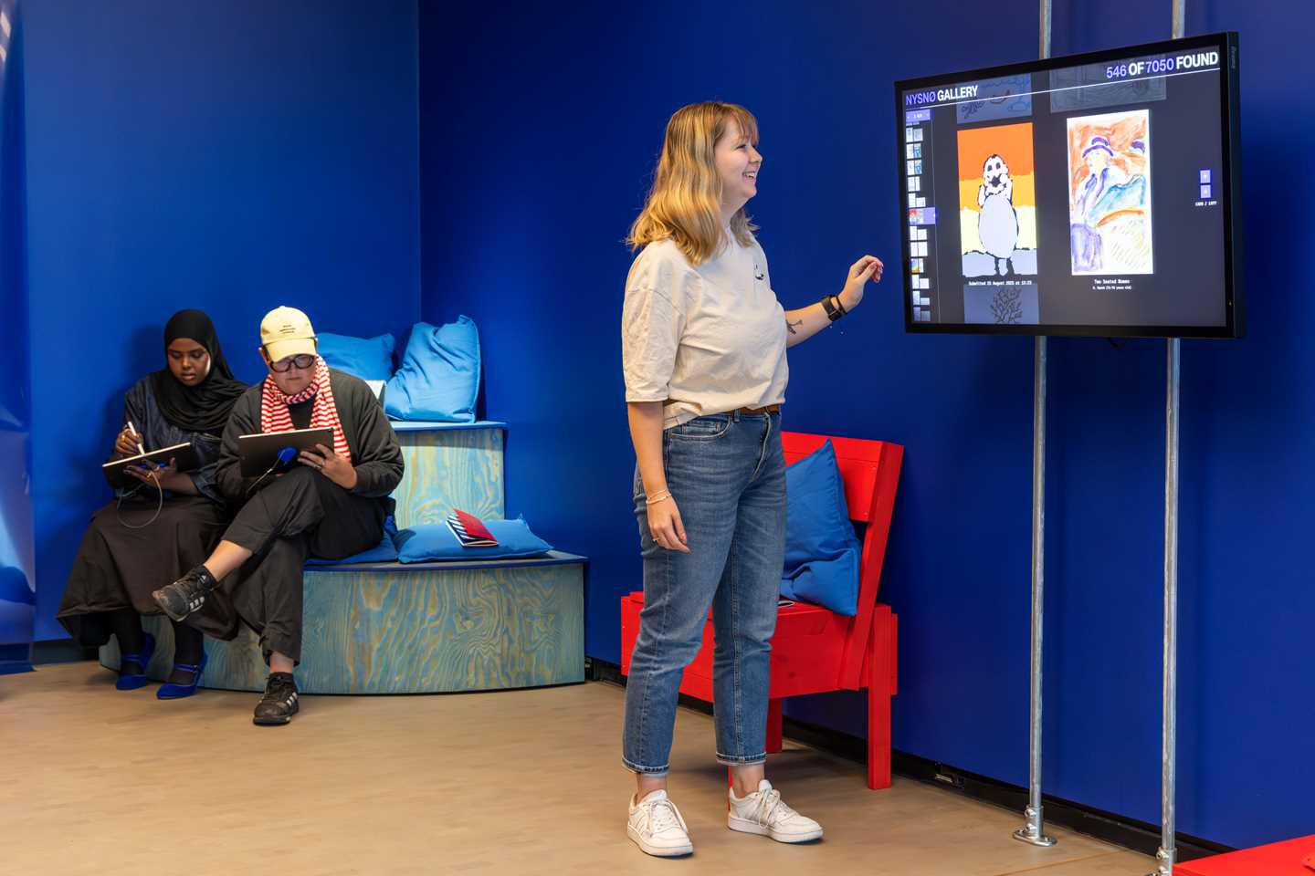 A girl scrolling through digitally submitted drawings on a screen. In the background, two people are sitting and drawing on tablets. Photograph.