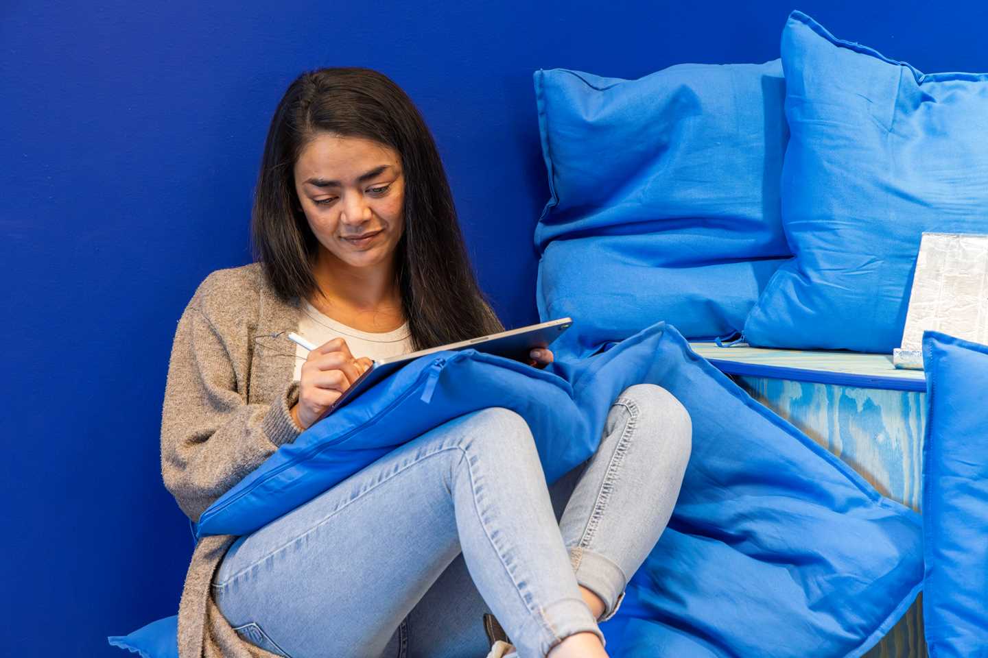 A woman sitting relaxed among many blue cushions, drawing on a tablet. Photograph.