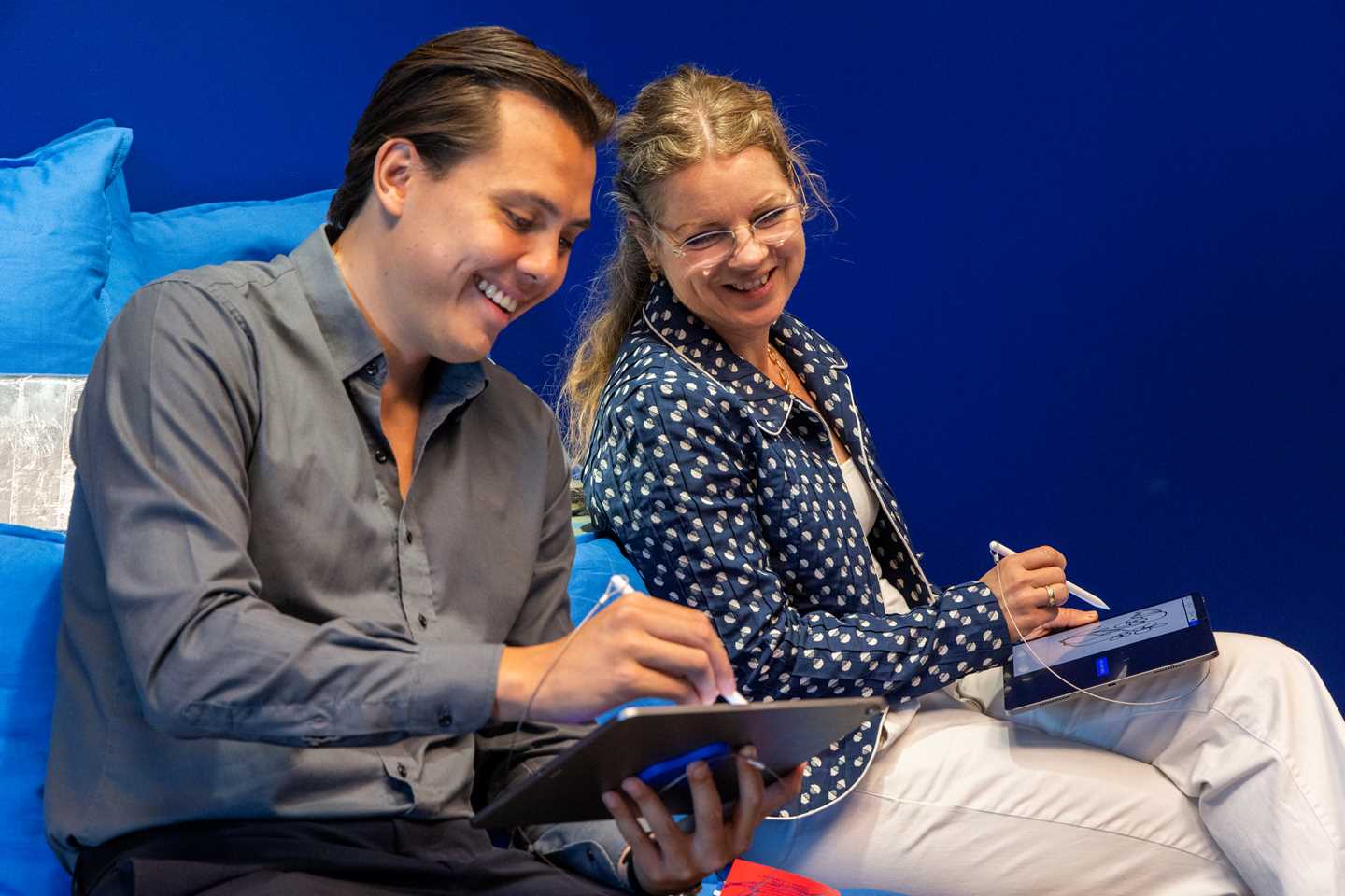 A man and a woman sitting and laughing as the man shows the woman what he has drawn on a tablet. Photograph.
