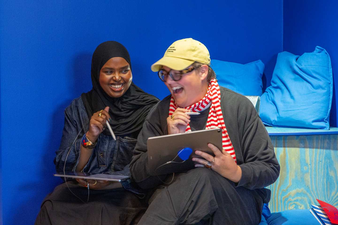Two girls sitting with their own tablets, laughing. Photograph.