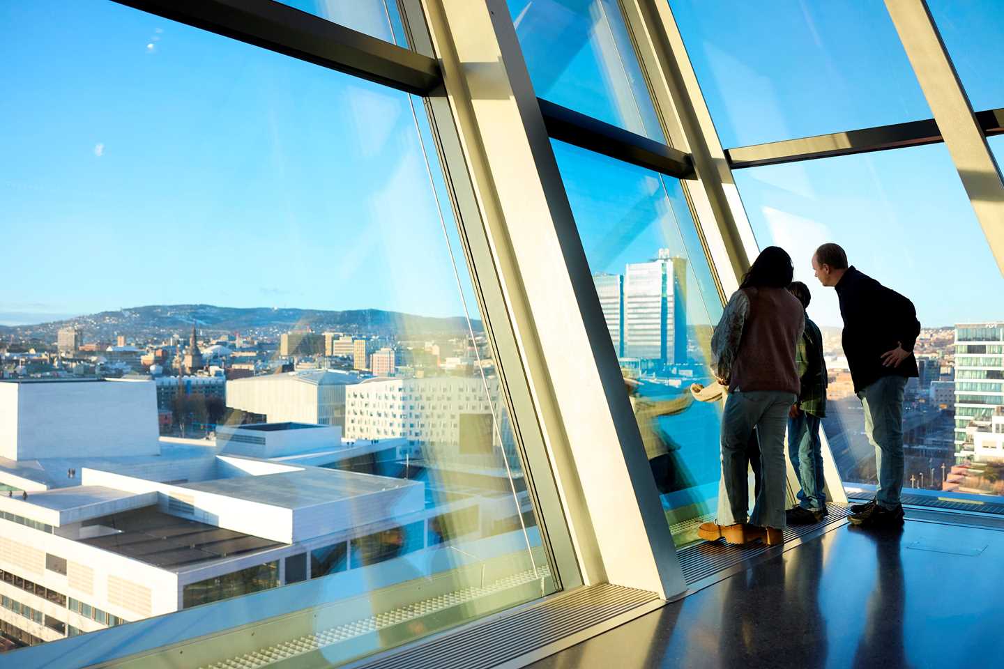 A family stands by the windows on the 10th floor of MUNCH, looking out over Oslo. Outside, the sky is blue and the Opera House is visible. Photograph.