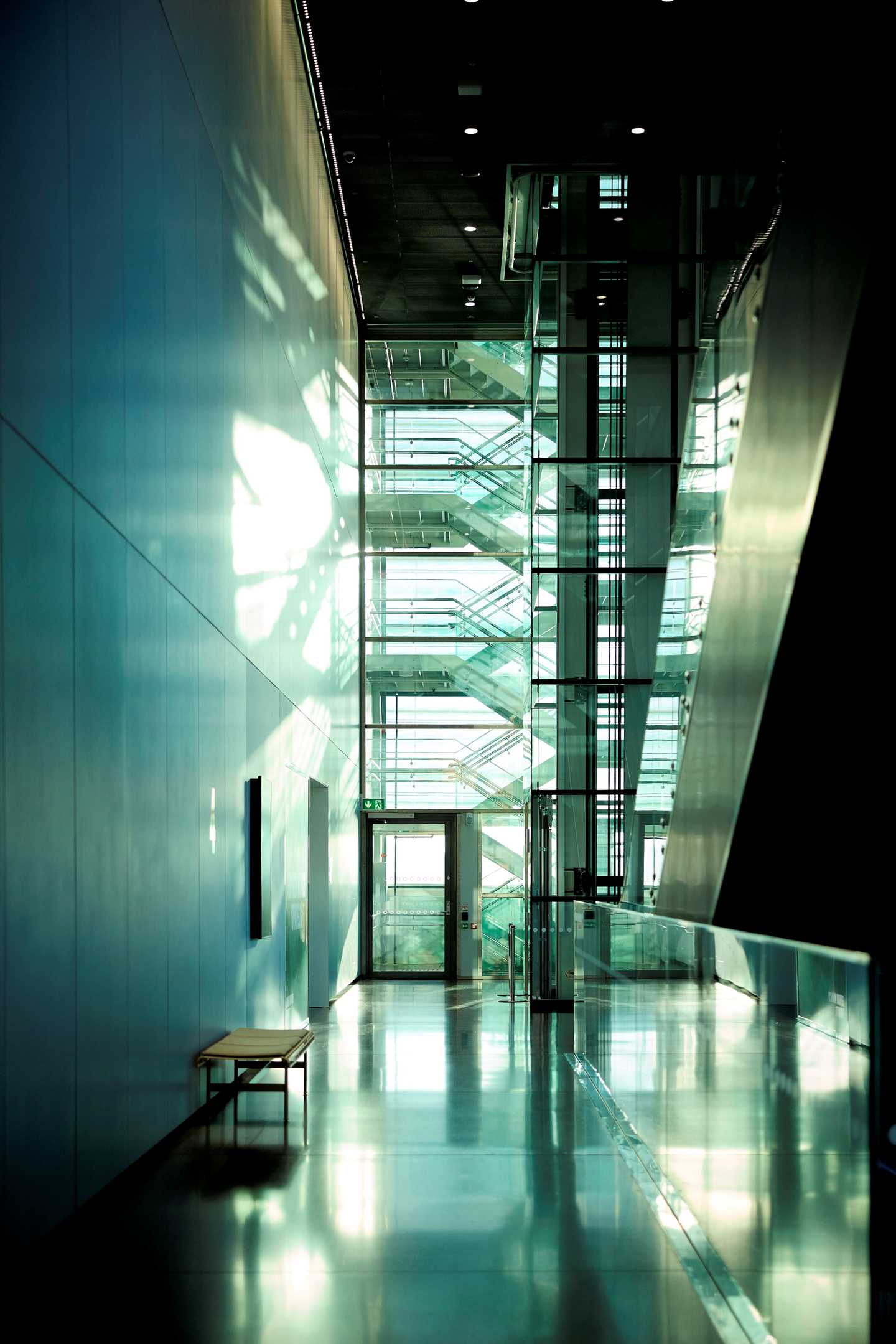 One of the corridors at MUNCH, with a bench on one side and an escalator on the other. At the end of the image, a staircase zigzags upwards. Photograph.