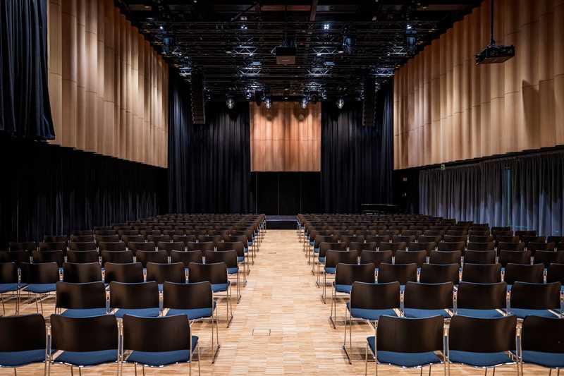 The banquet hall at MUNCH seen from the back. Several rows of black chairs face a stage. Photograph.