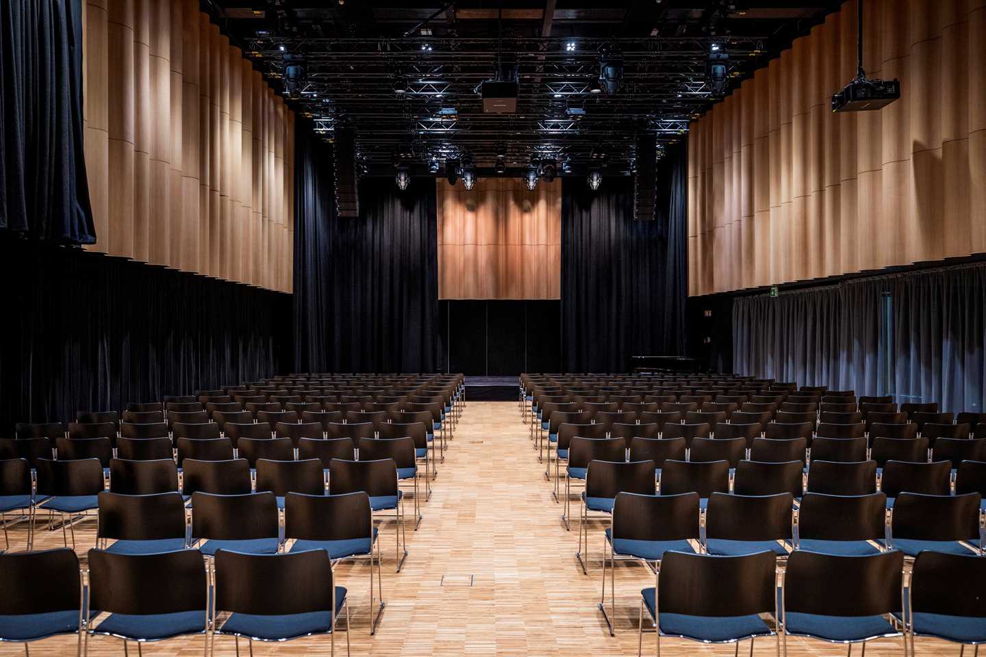 The banquet hall at MUNCH seen from the back. Several rows of black chairs face a stage. Photograph.