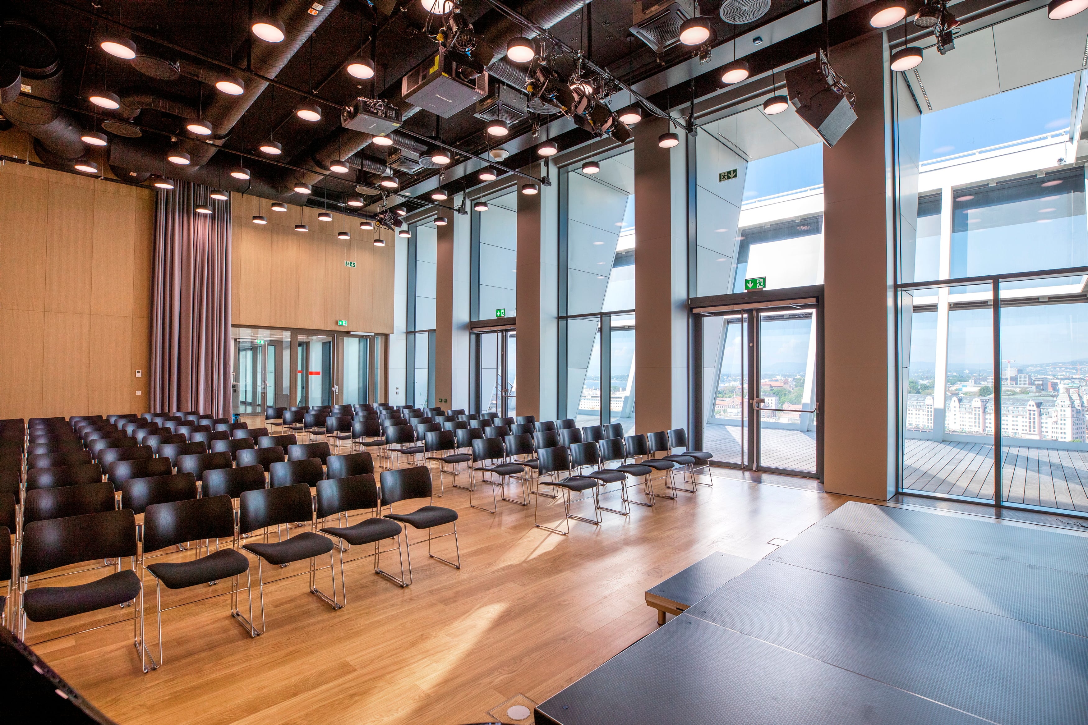 The top hall at MUNCH seen from the stage. Inside the room are rows of black chairs, and one wall consists of floor-to-ceiling windows, with a rooftop terrace visible outside. Photograph.