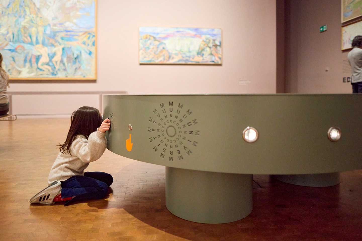 A young girl kneels, looking through a peephole in an installation in the Monumental exhibition.