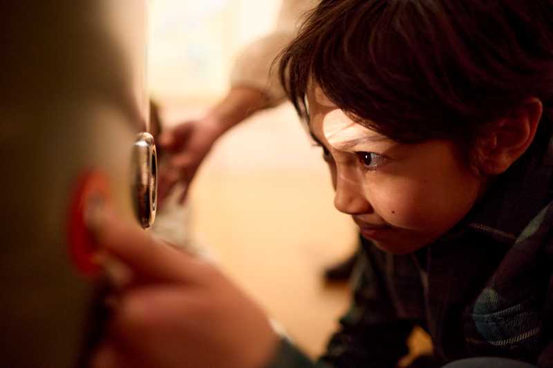 A young boy crouches, staring into a peephole in an installation in the Monumental exhibition. Photograph.
