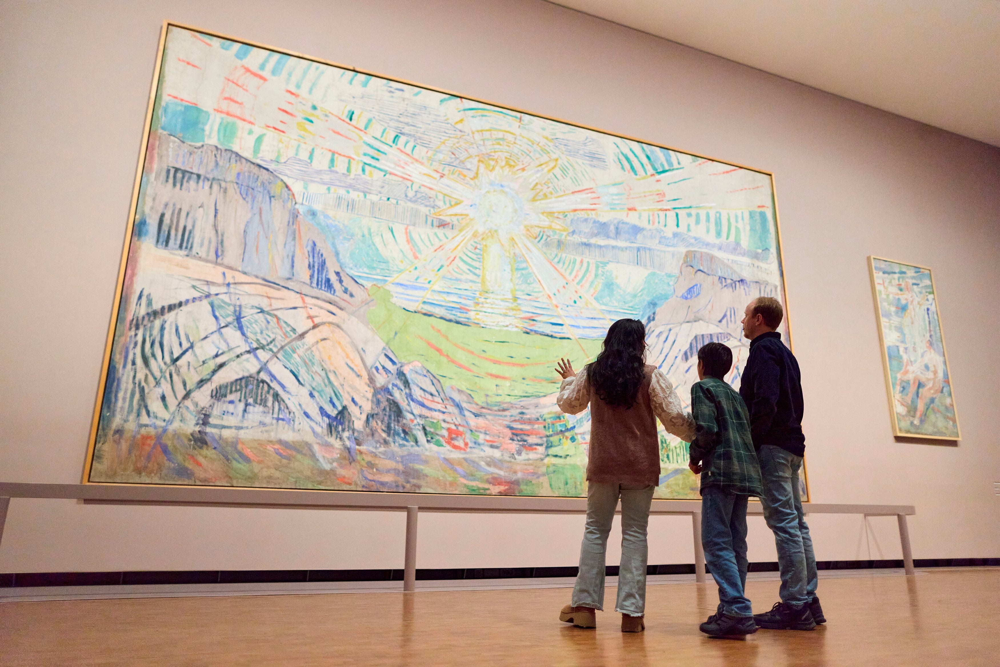 A mother, son and father stand looking at The Sun by Edvard Munch. They are facing away from the camera. Photograph.