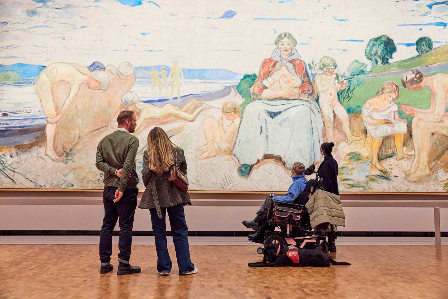 Four people stand with their backs to the camera, observing the painting Alma Mater by Edvard Munch. The artwork is 50 square metres in size. Photograph.