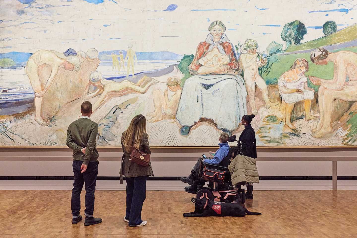 Four people stand with their backs to the camera, observing the painting Alma Mater by Edvard Munch. The artwork is 50 square metres in size. Photograph.