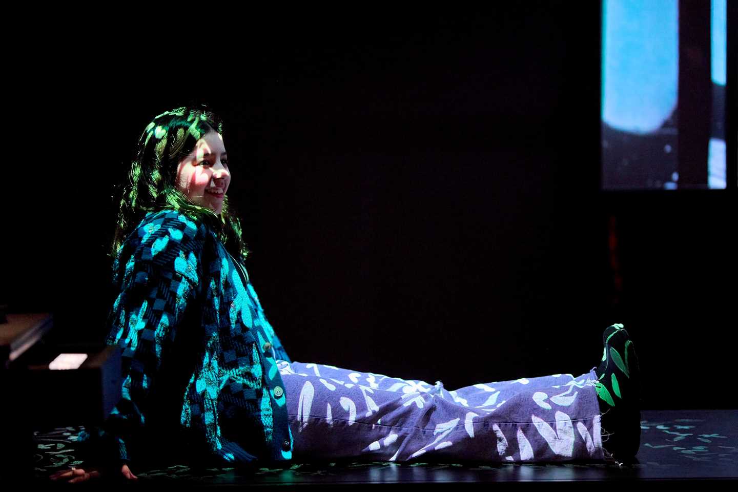 A girl sits on a bench, both she and the bench illuminated by glowing flower drawings projected from the ceiling. Photograph.