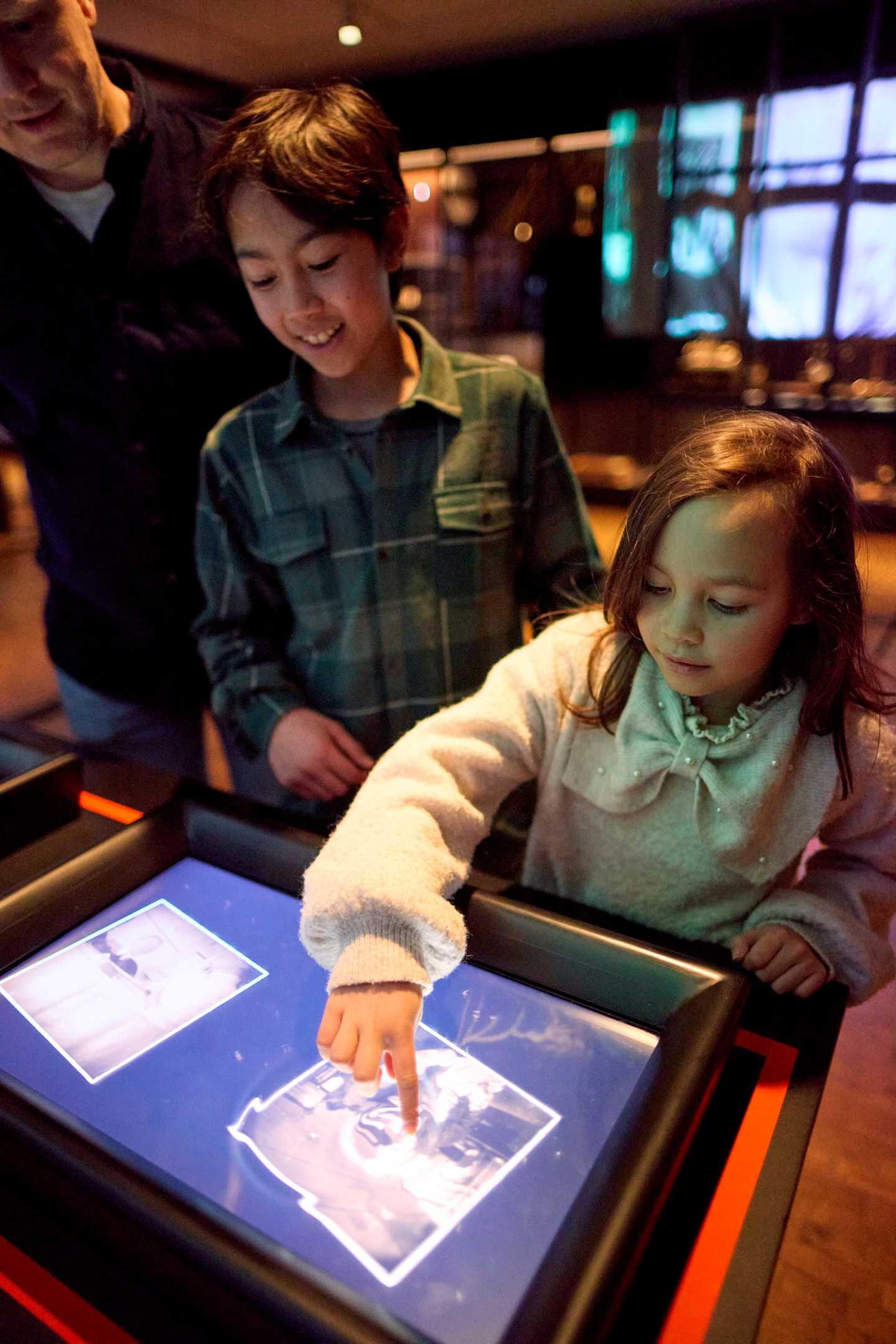 Two children and a father touch a digital screen in the Shadows exhibition. Photograph.