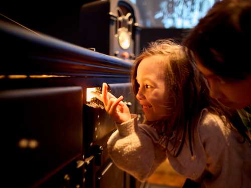 Two children crouch and look through a mouse hole with light coming out of it. Photograph.