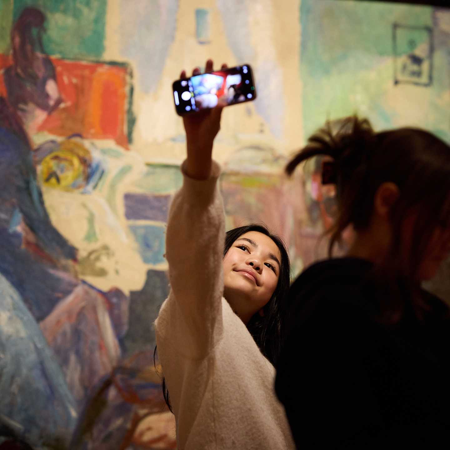 En ung jente tar et bilde av seg selv med mobilen sin foran et maleri av Edvard Munch i utstillingen Uendelig. Fotografi