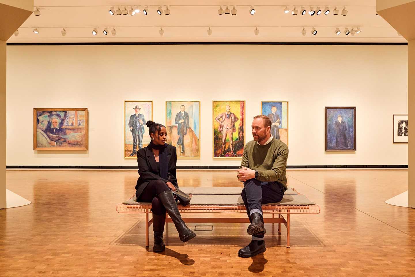 Man and woman talking on a bench in front of Munch portraits. Photograph