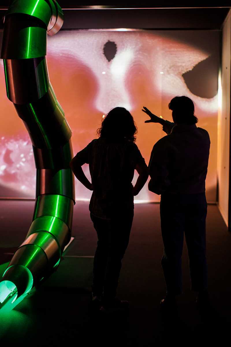 Two people stand in an exhibition hall, viewing artwork by Sandra Mujinga. They are seen from behind. Photograph.