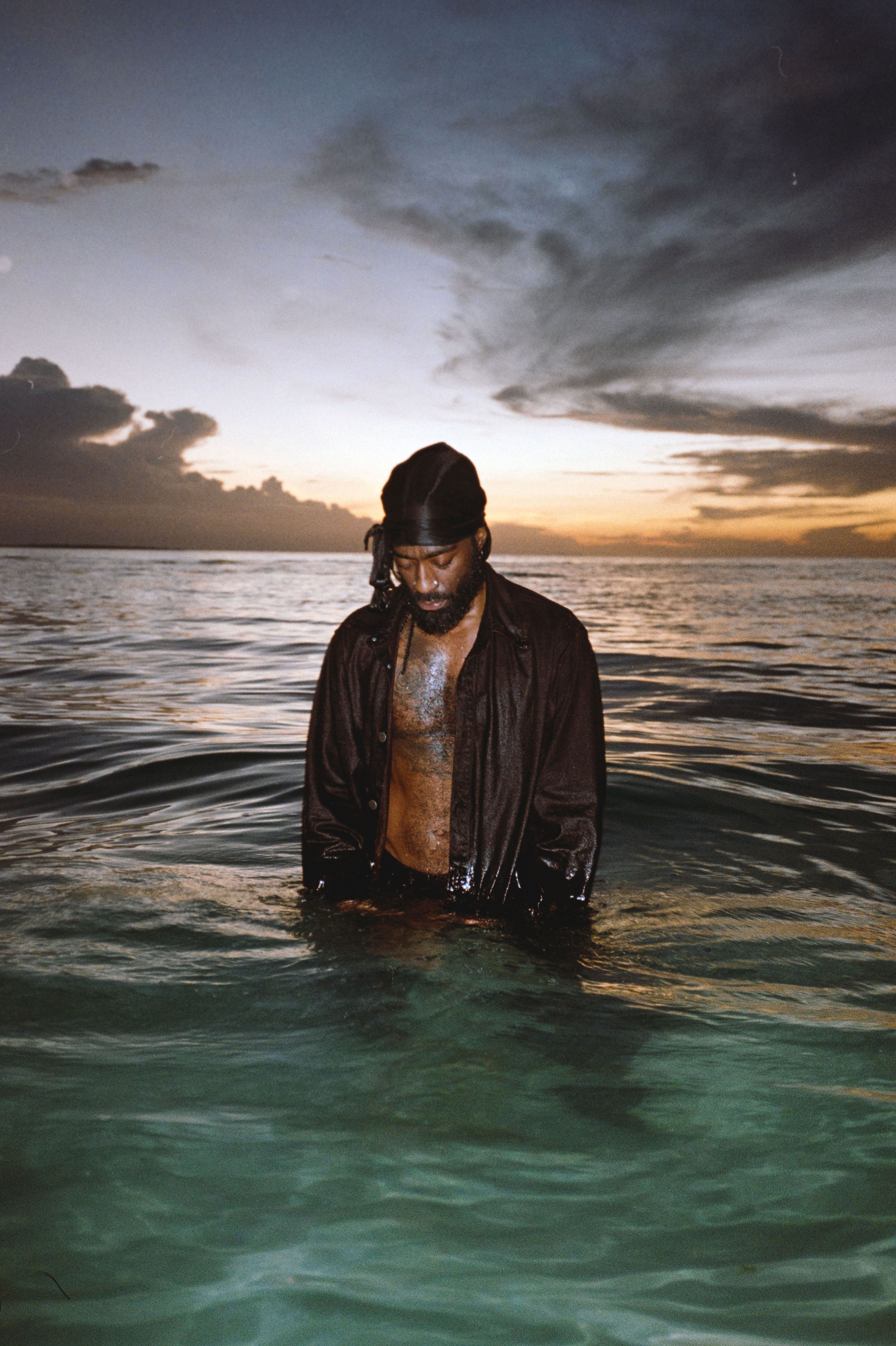 Arif stands in the sea with water up to his thighs, looking down into the water. He is wearing an open black shirt, and there is a sunset in the background. Photograph.