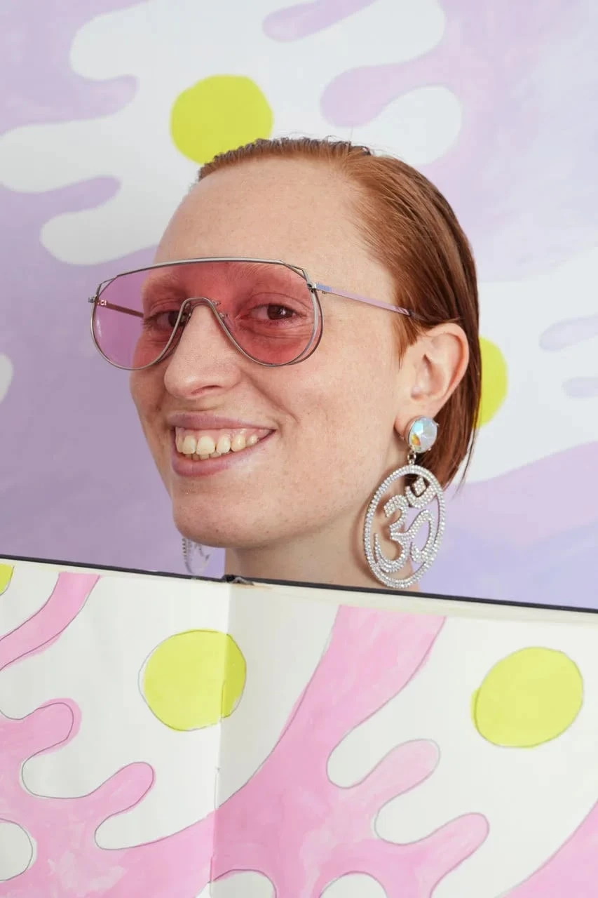 Constance Tenvik wearing pink sunglasses, smiling with a sketchbook. Photograph.