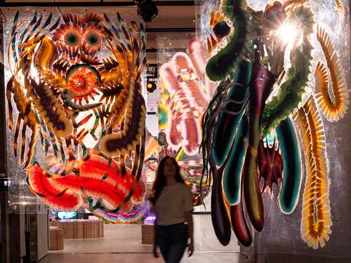 A woman walks through a corridor where paintings hang down from the ceiling. Photo