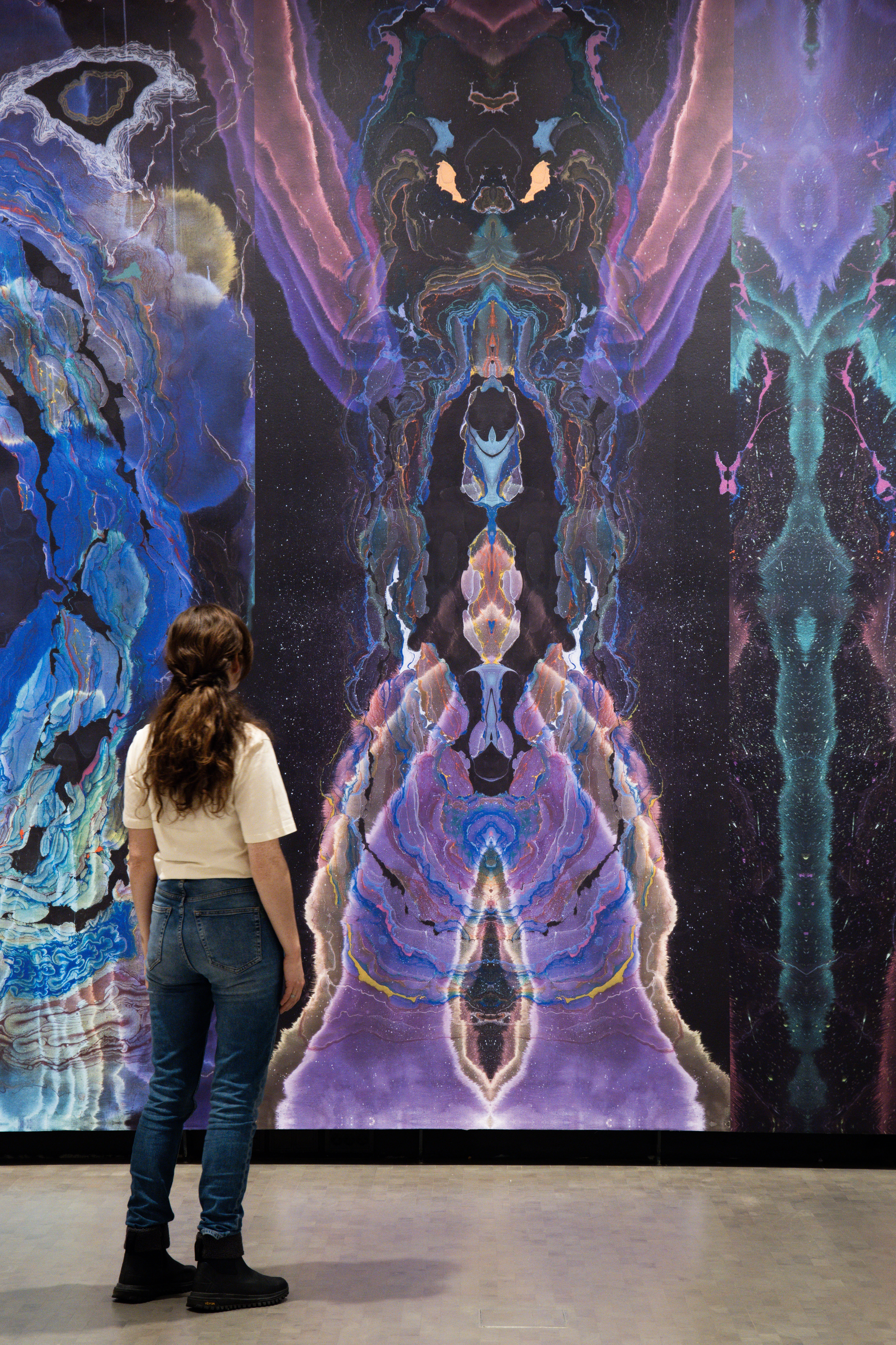 A woman stands with her back turned, observing a wall covered in wallpaper created by artist Kerstin Brätsch. Photograph.