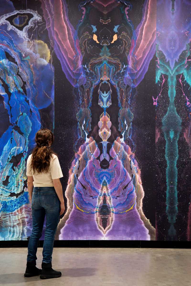 A woman stands with her back turned, observing a wall covered in wallpaper created by artist Kerstin Brätsch. Photograph.