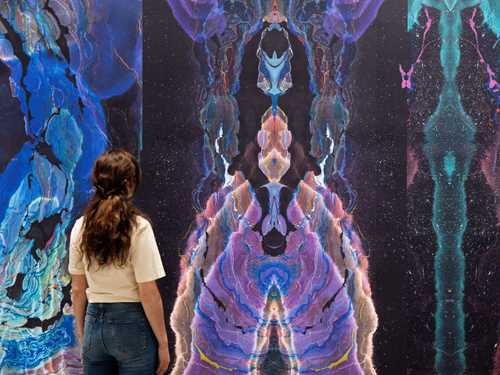 A woman stands with her back turned, observing a wall covered in wallpaper created by artist Kerstin Brätsch. Photograph.