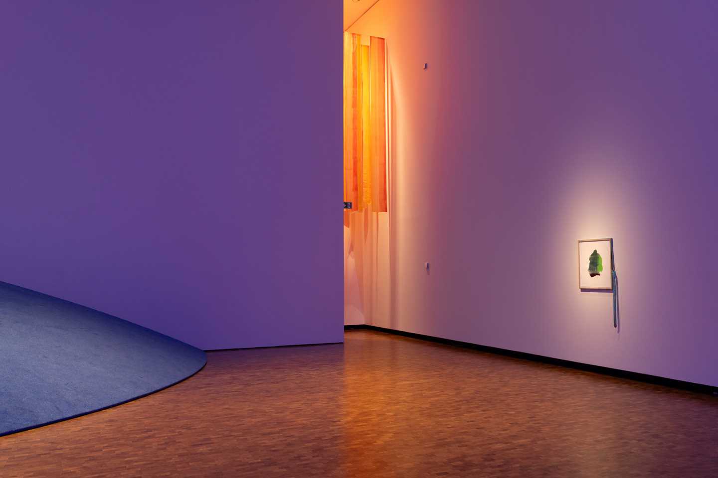 Parts of the exhibition space display a stunning interplay of blue, purple, orange, and yellow. A piece of artwork hangs on one wall. Photograph.