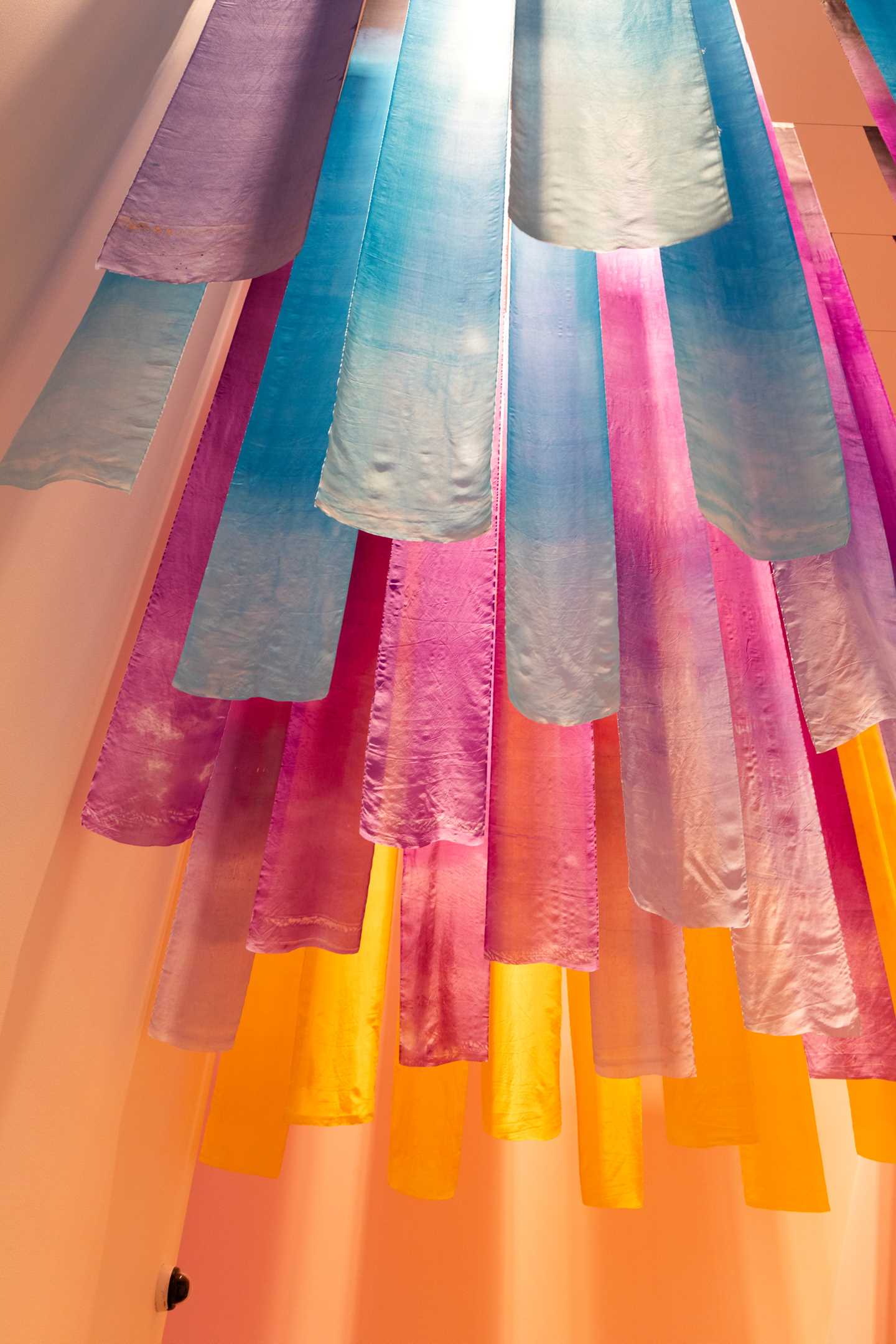 Thick strips of silk fabric in various colours hang from the ceiling, leading in and out of the exhibition. Photograph.