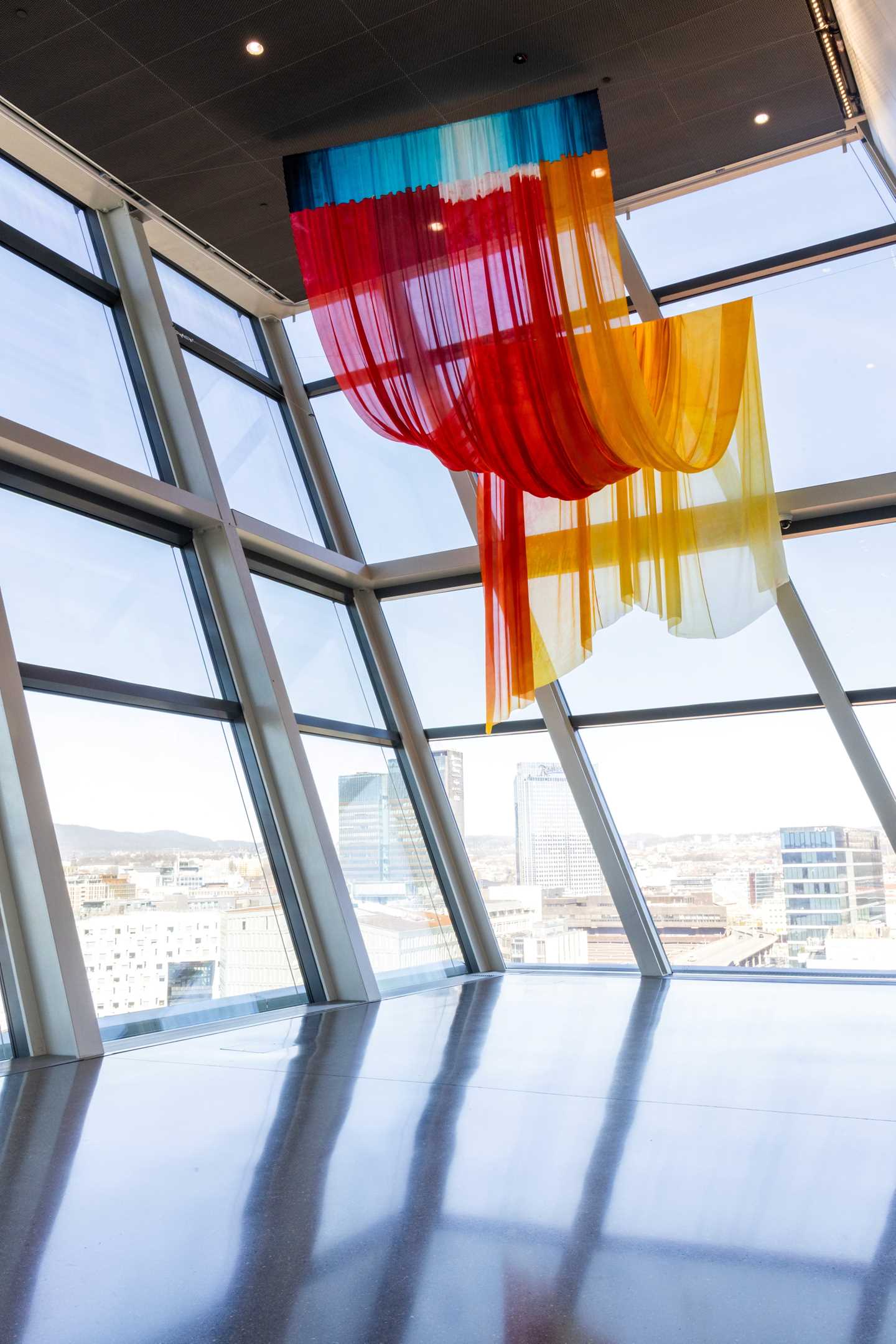 A large silk fabric in various colours hangs in waves from the ceiling. In the background, windows reveal a view of Oslo city. Photograph.