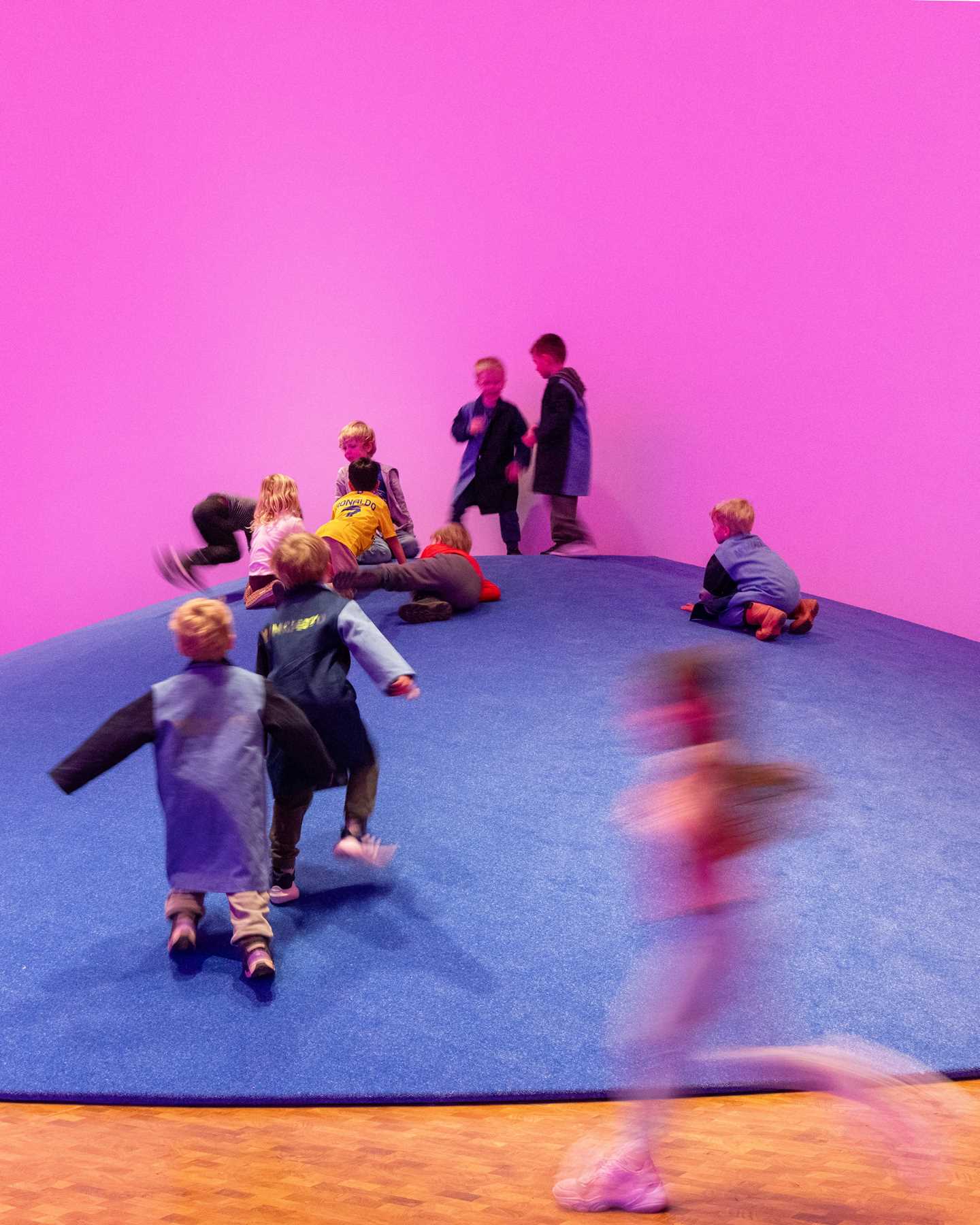 Many children enjoy running and climbing on a blue amphitheatre that forms a mountain-like structure in the exhibition space. Photograph.