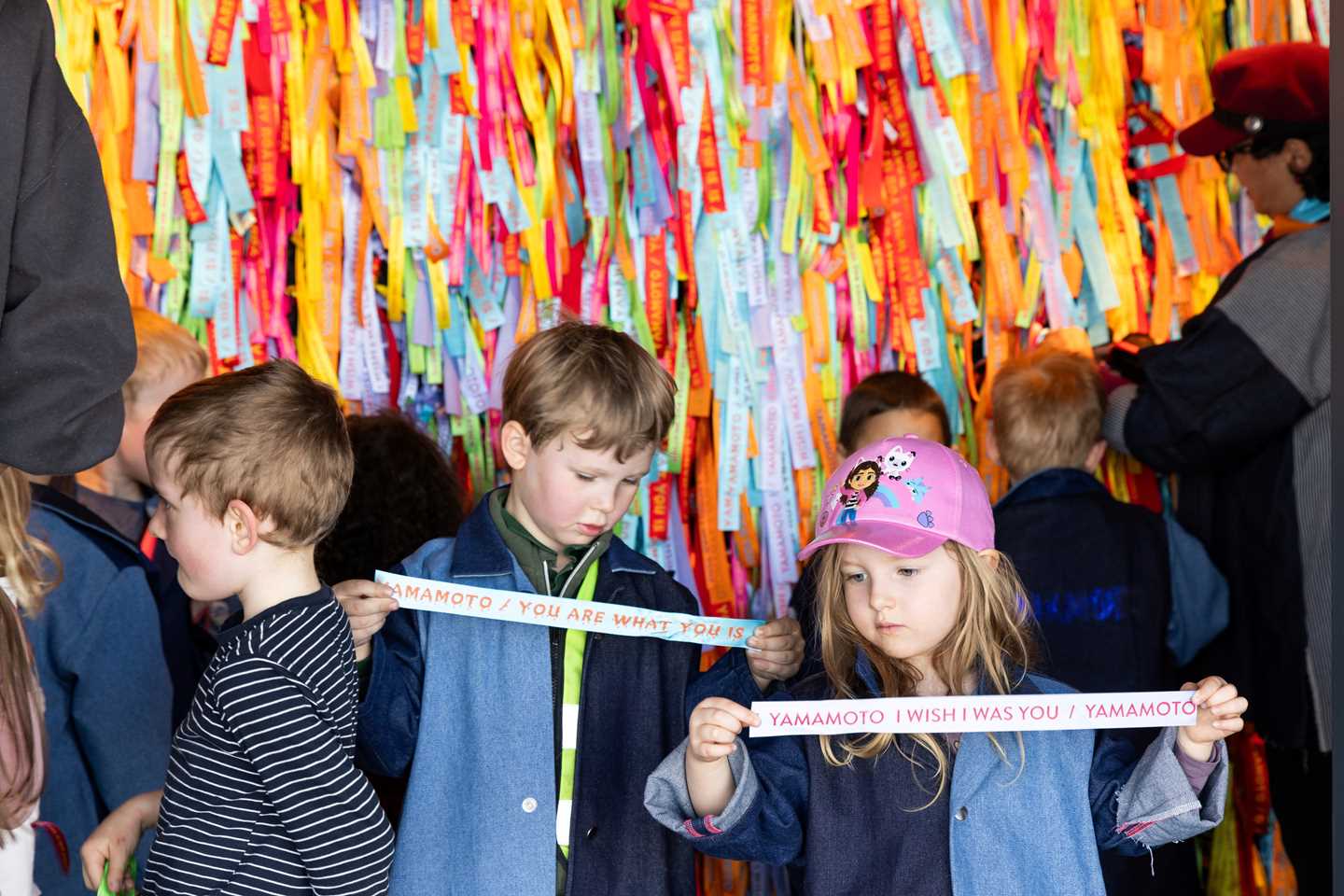 Children stand in front of the wall of Brazilian ribbons, each holding one and examining it closely. Photograph.