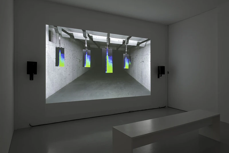 A small white room with a projection on the wall showing sound frequencies. On each side of the projection there is a black speaker, and on the floor in front of the projection there is a white bench. Photography
