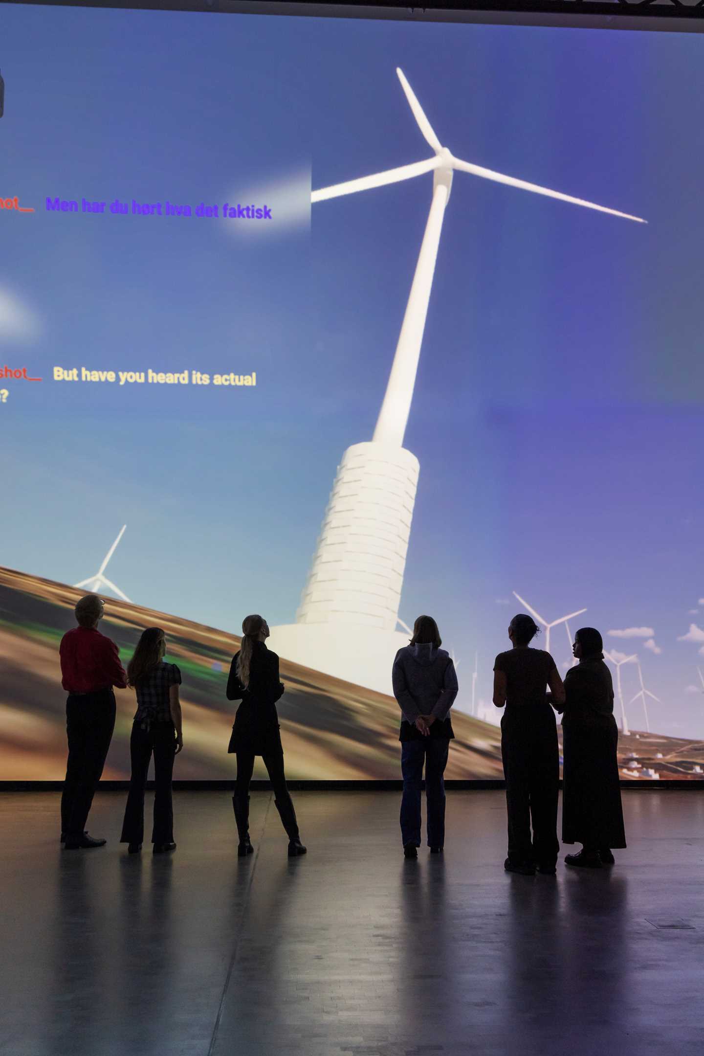 A group of people stand in a dark room in front of a wall covered by a projection showing a computer animation of wind turbines on the Golan Heights, along with a box displaying a chat dialogue. Photograph
