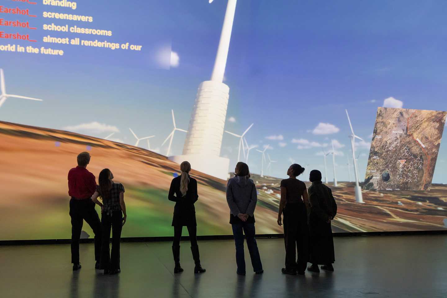 A group of people stand in a dark room in front of a wall covered by a projection showing a computer animation of wind turbines on the Golan Heights. Photograph