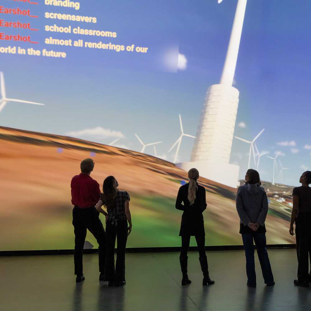 A group of people stand in a dark room in front of a wall covered by a projection showing a computer animation of wind turbines on the Golan Heights. Photograph